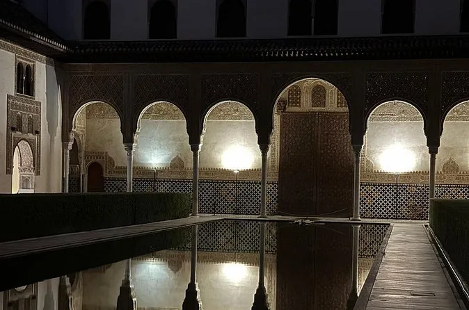 A lit arcade of the Court of the Myrtles reflected in dark water at night